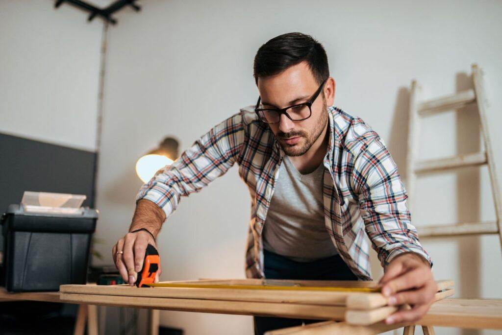 Ein Heimwerker misst Holzlatten aus – sorgfältige Planung und handwerkliche Präzision für den Bau eines funktionalen Holzlagers.